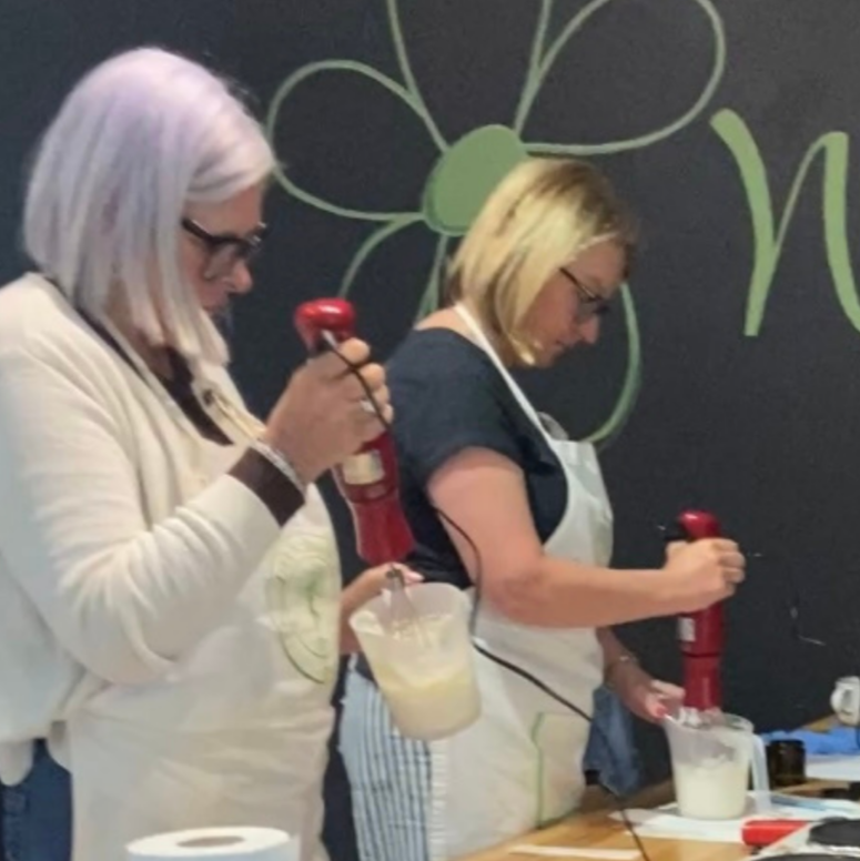 Two women standing at a wooden table, each holding a red handheld electric mixer and pouring a pale liquid into a measuring cup. The scene is indoors against a dark wall with a large green flower outline.