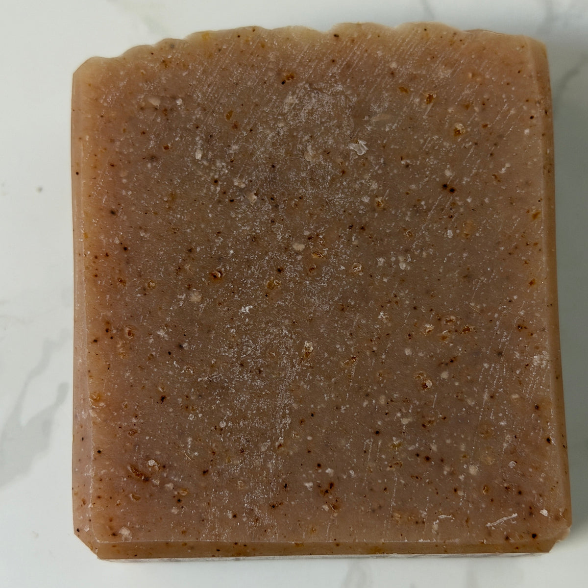 A rectangular bar of brown soap with coarse speckled exfoliating particles, photographed from directly above on a white marble surface.