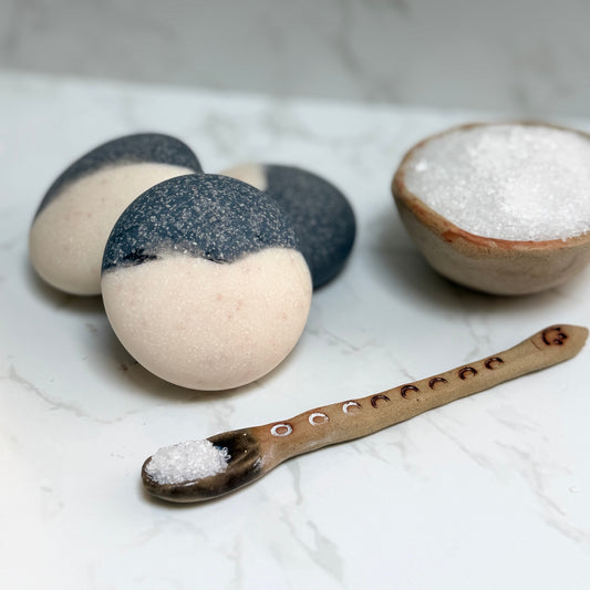 Three oval spa salt bars with a cream and black split design are arranged on a white marble surface next to a small ceramic bowl of white hymalyan salt and a wooden spoon heaped with salt