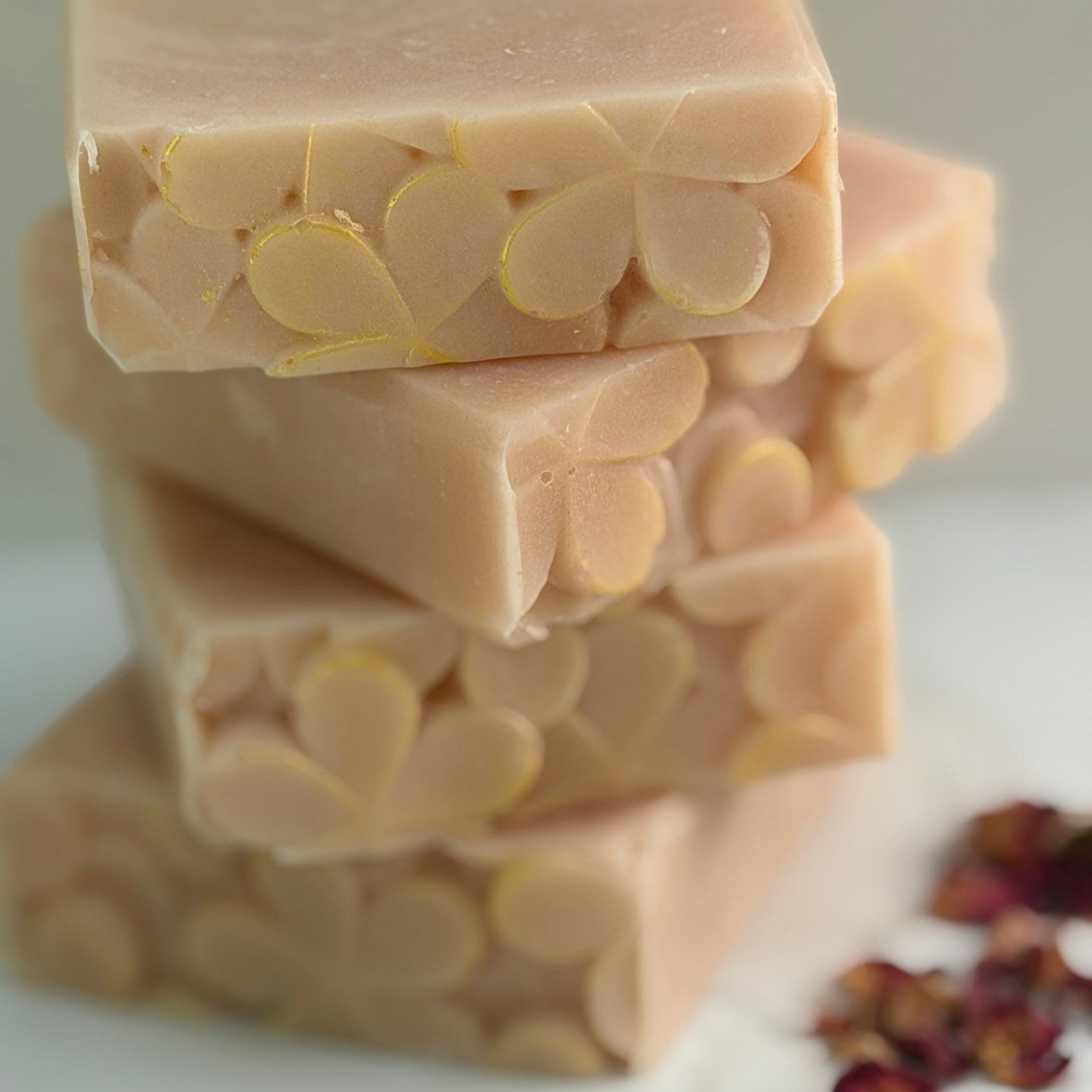 A zoomed shot of stacked bars of pale pink-colored handmade soap with a carved floral pattern, set on a light marble surface with scattered dried rose petals.