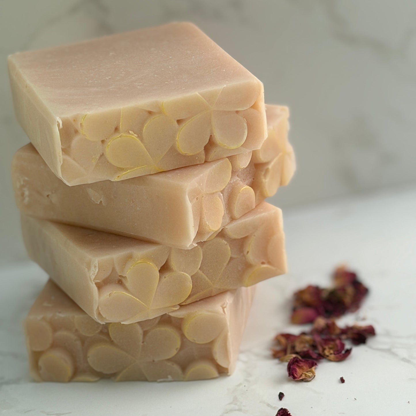Three stacked hand made rectangular bars of pale pink colored soap with visible floral imprint, arranged on a light marble surface with scattered dried rose petals.