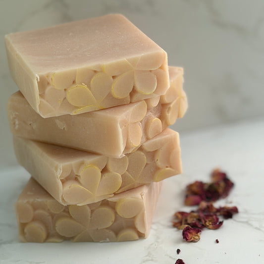 Three stacked hand made rectangular bars of pale pink colored soap with visible floral imprint, arranged on a light marble surface with scattered dried rose petals.