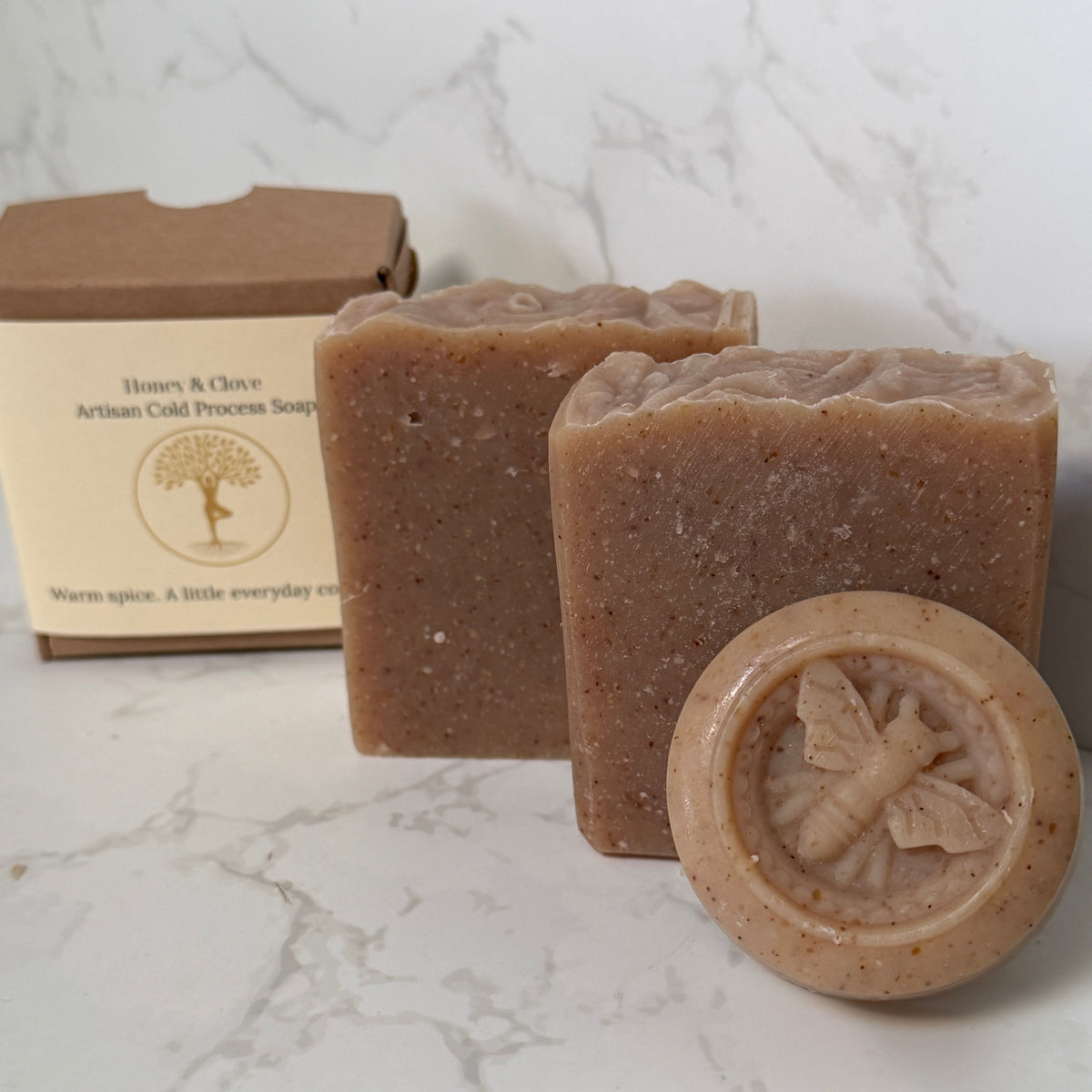 Two rectangular bars of honey spice artisan cold process soap and one round stamped bar in front, displayed next to their kraft paper packaging box on a white marble surface.