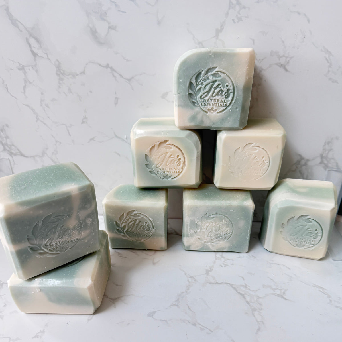 A group of handmade rectangular soap bars in a pale sage green and cream marbled pattern, arranged on a white marble surface against a matching marble background. The soaps feature an embossed circular logo on the top face.