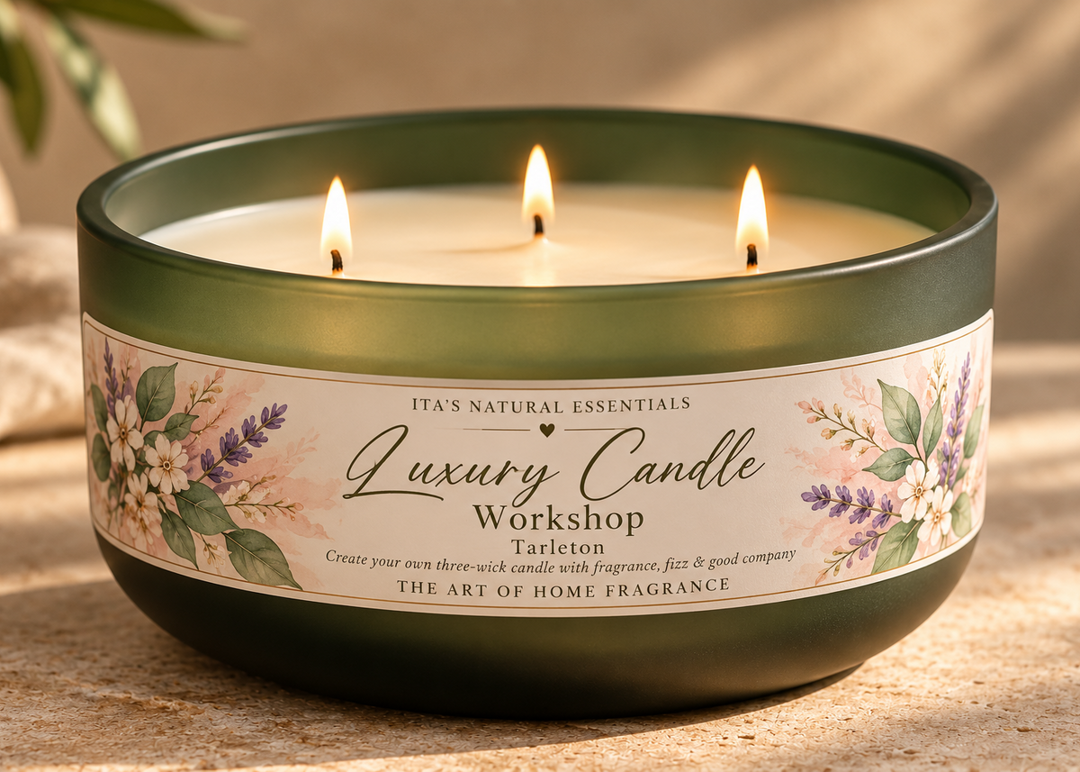 A three-wick scented candle in a dark green glass jar with a floral label reading 'Luxury Candle Workshop Tarleton' and 'It's Natural Essentials,' shown lit on a stone surface with warm sunlight and soft background blur.